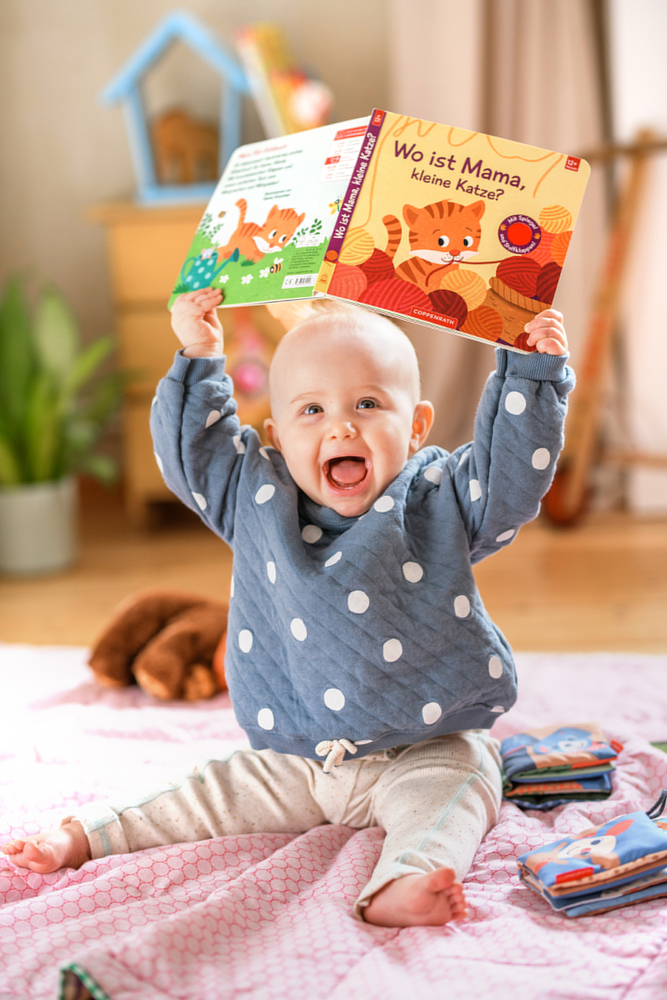 Ein lächelndes Baby sitzt drinnen auf einer Decke und hält ein buntes Bilderbuch hoch.