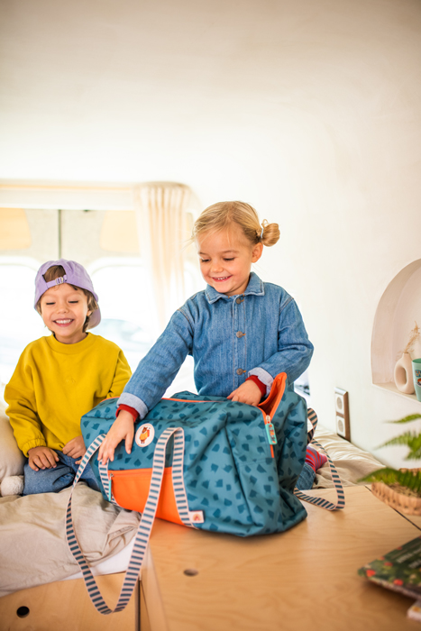 Banner zum Thema Reise: Ein Mädchen und ein Junge in einem Wohnmobil öffnen eine Kinderreisetasche und lachen.