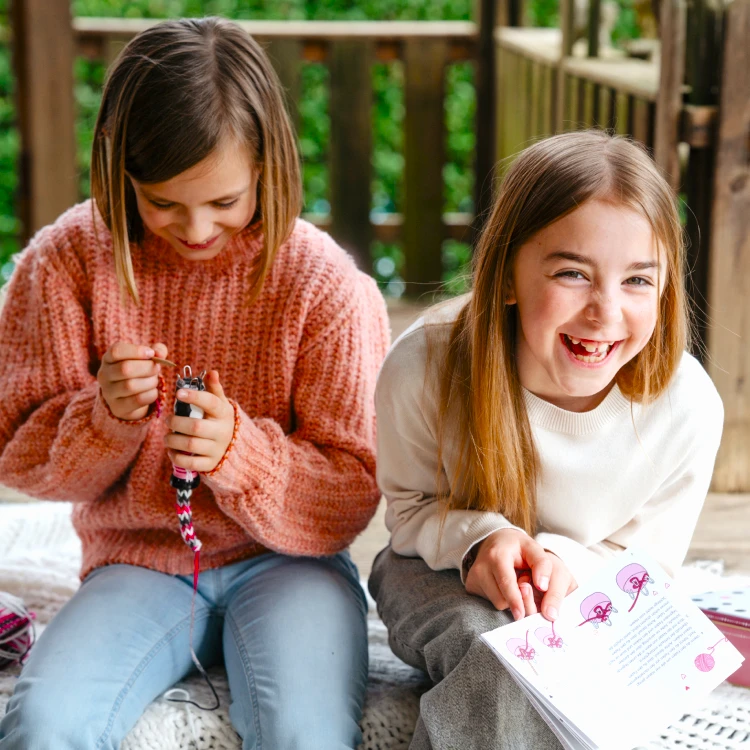 Zwei lachende Mädchen auf einer Terrasse machen Handarbeiten.