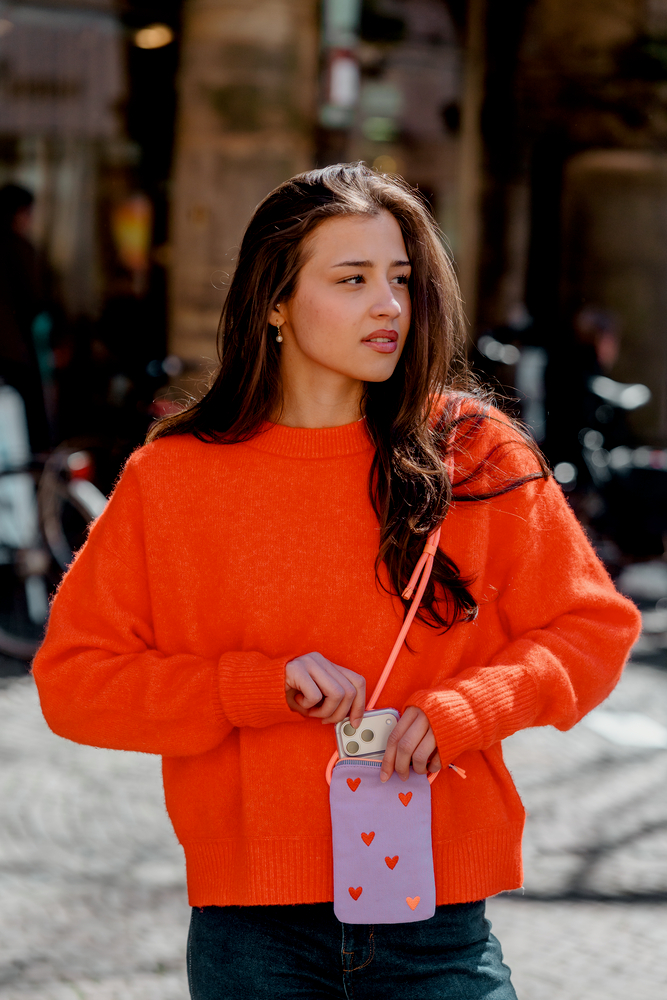 Person mit orangefarbenem Pullover hält eine lilafarbene Handytasche mit roten Herzen und einem sichtbaren Smartphone.