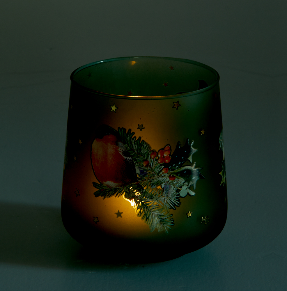 Eine Kerze leuchtet in einem festlichen Windlicht, das mit Stechpalmen, roten Beeren und goldenen Sternen geschmückt ist.