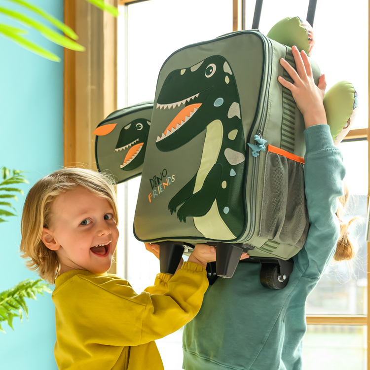 Kleiner Junge lacht in die Kamera. Daneben ein Kind, das einen grünen Trolley mit Dino-Motiv in die Höhe hält.