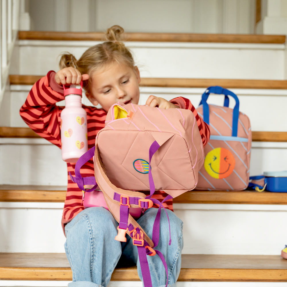 Junges Mädchen sitzt auf Treppe, hält eine Wasserflasche und schaut in ihren pinken Rucksack.