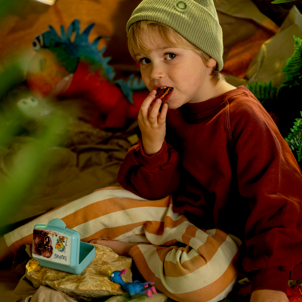 Kind mit gestreifter Hose und Mütze sitzt und isst Snack, umgeben von Spielzeugdinosauriern und Pflanzen.