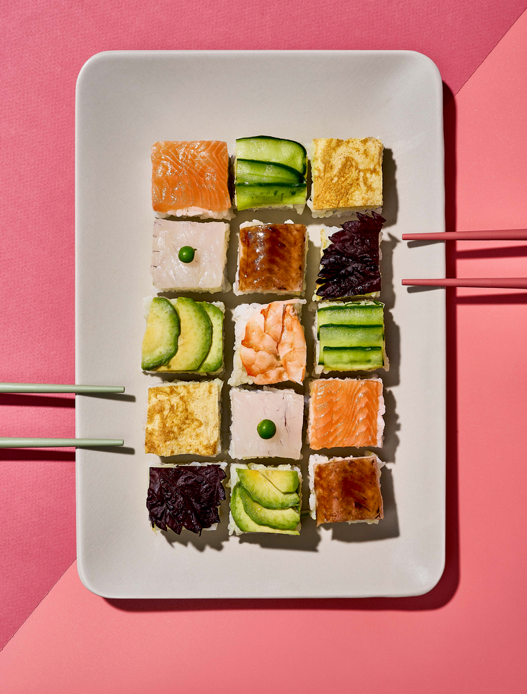 Rechteckiger Teller mit 15 bunten Sushi-Würfeln, gitterförmig angeordnet, vor rosa Hintergrund und mit rosa Stäbchen.