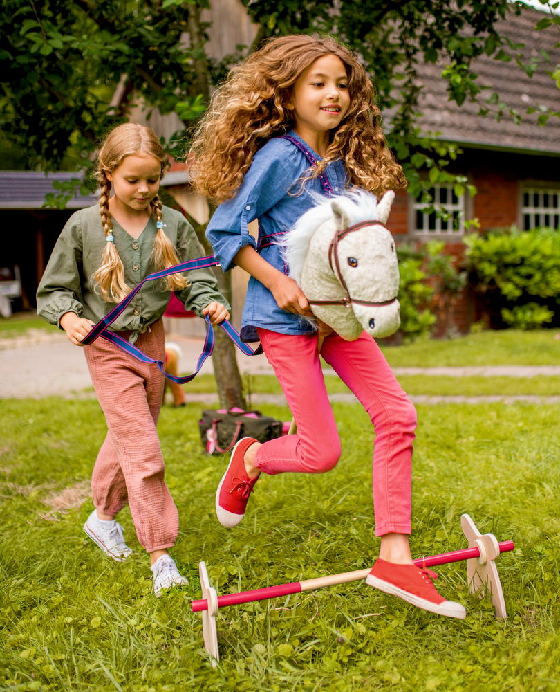 Zwei Mädchen spielen draußen: Eines springt mit einem Steckenpferd, das andere folgt mit Bändern.