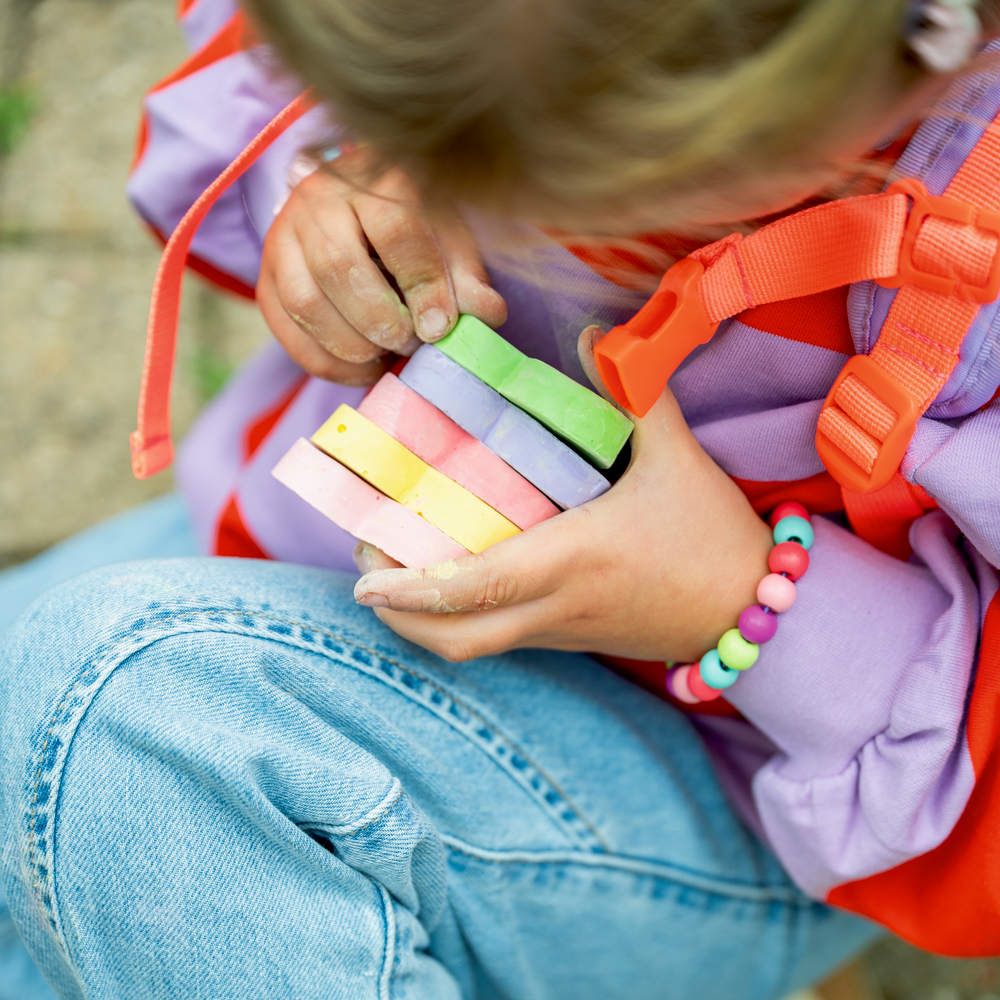 Kind in bunter Kleidung sitzt am Boden und hält Pastellkreide, Aufnahme von oben.