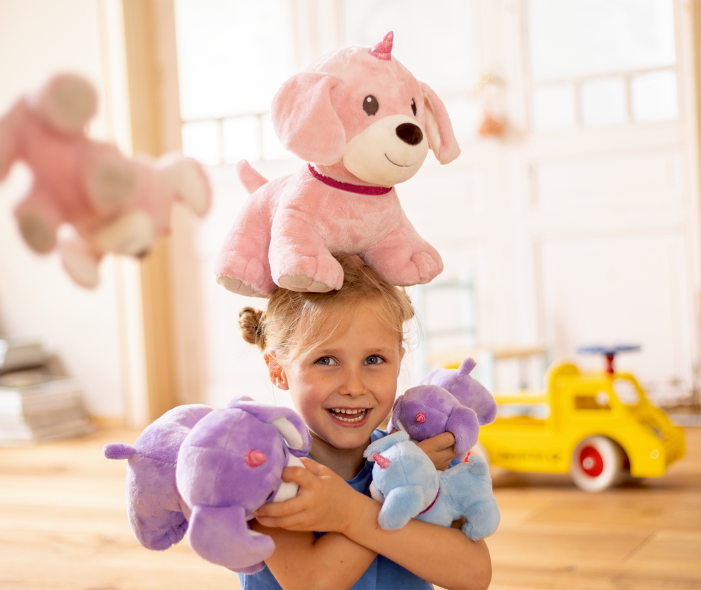 Lächelndes Mädchen im sonnigen Spielzimmer balanciert rosa Plüschhund auf dem Kopf und hält Stofftiere in der Hand.