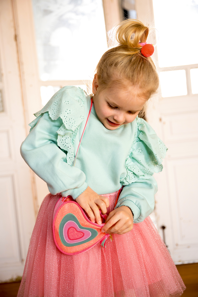 Kind mit mintgrünem Oberteil und rosa Tüllrock hält eine herzförmige Tasche in Pink- und Türkistönen.