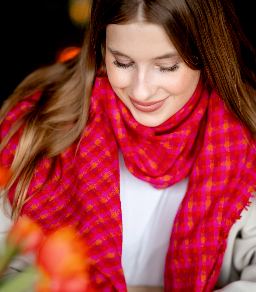 Lächelnde junge Frau mit langen braunen Haaren und einem rot-pink karierten Schal, die nach unten blickt.