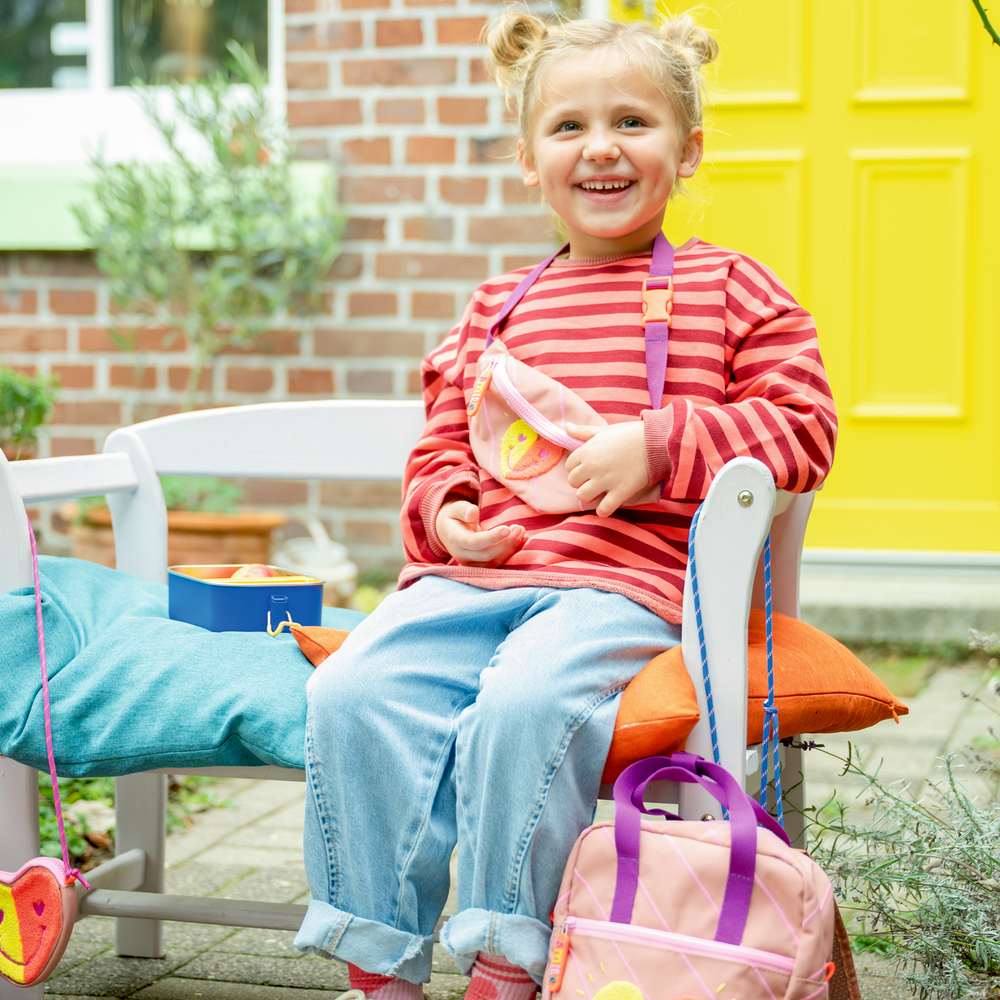 Junges Mädchen mit gestreiftem Shirt sitzt auf Bank. Daneben Rucksack, Brotdose. Im Hintergrund gelbe Tür.