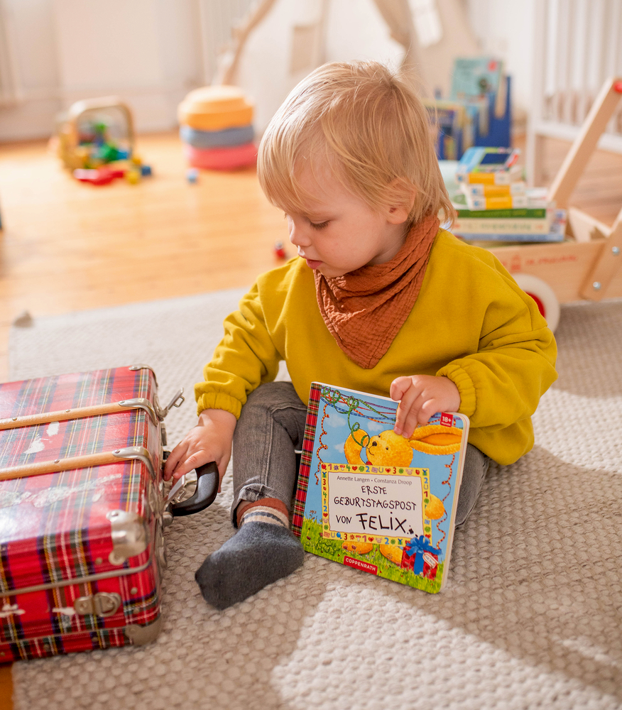Kleinkind im gelben Pullover sitzt auf dem Boden, hält ein Buch und berührt einen rot karierten Koffer.