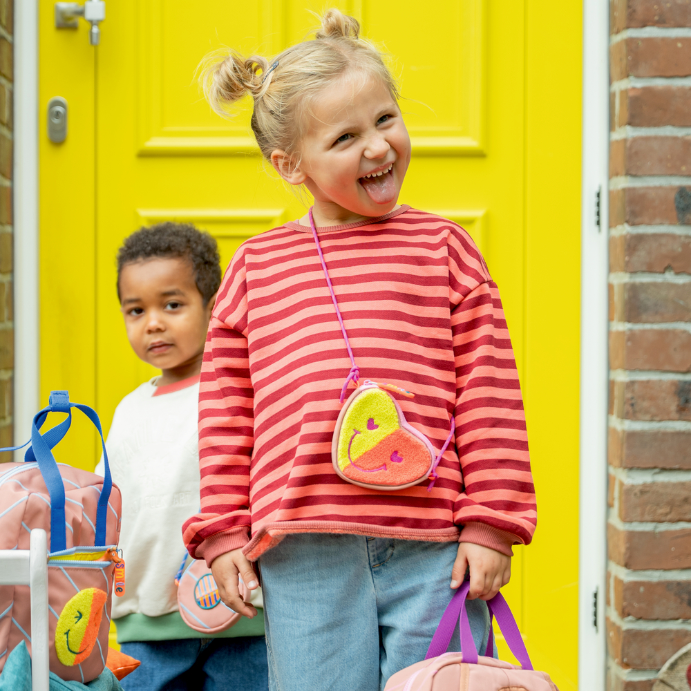 Ein Mädchen im rot gestreiften Shirt steht vor einer gelben Tür, ein Junge und Taschen sind daneben.