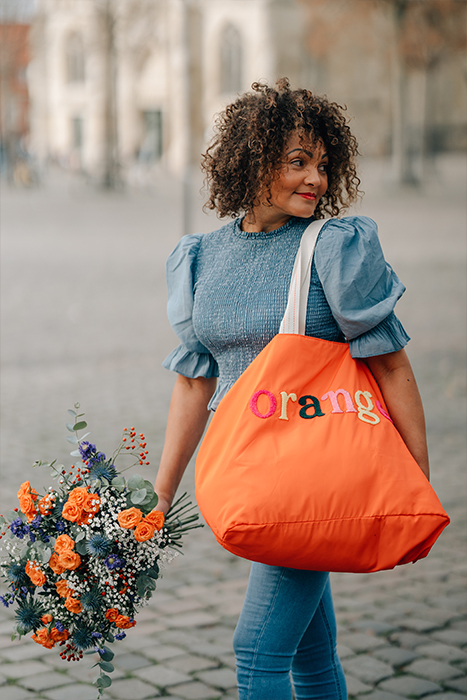 Banner zum Thema Taschen & Shopper: Eine Frau mit Locken und einem Blumenstrauß schlendert mit einem großen orangefarbenen Shopper über den Münsteraner Domplatz 