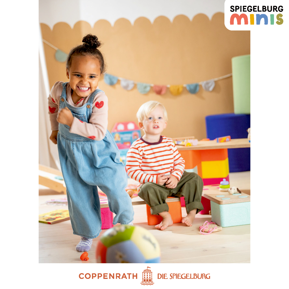 Ein Mädchen lächelt und läuft auf einen Ball zu. Ein Junge sitzt daneben und schaut zu. Im Hintergrund ist ein heller Raum.