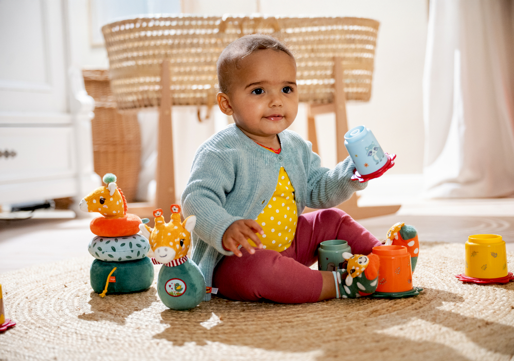Ein Baby sitzt auf einem Teppich und spielt mit Stapelbechern und Plüschtieren in einem hellen Raum.