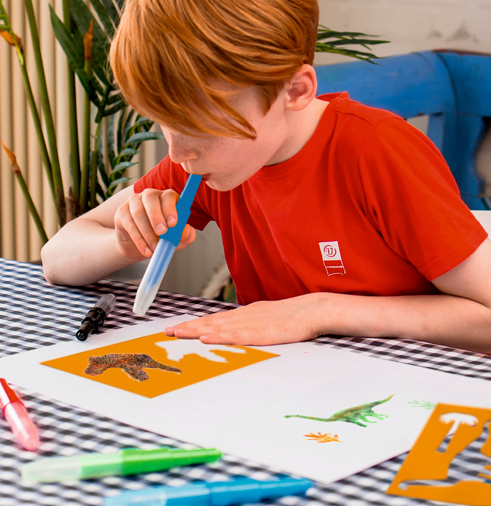 Ein rothaariges Kind pustet Farbe auf eine Schablone über einem weißen Blatt Papier.