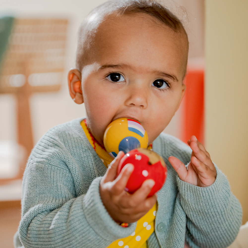 Baby in grünem Pullover hält buntes Spielzeug und kaut darauf.