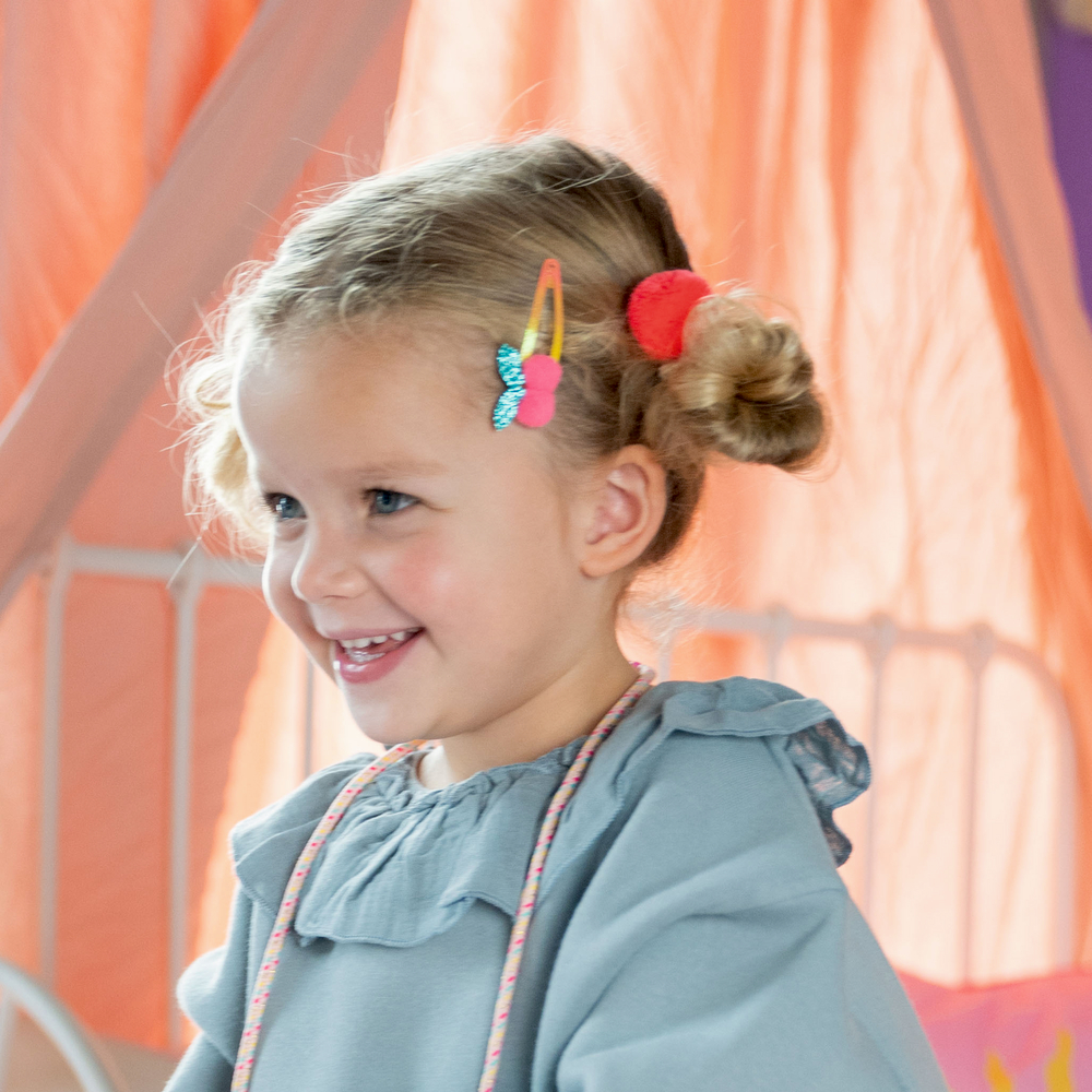 Junges Mädchen mit Zöpfen und bunten Haarspangen trägt ein hellblaues Oberteil mit Rüschen. Sie lächelt.