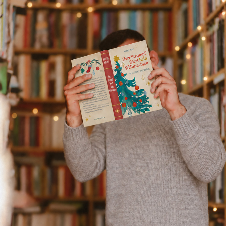 Mann vor Bücherrgal mit Lichterketten hält sich ein weihnachtliches buch vors Gesicht.