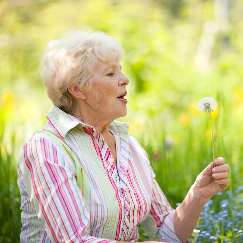 Ältere Frau bei einer Blumenwiese mit Pusteblume.