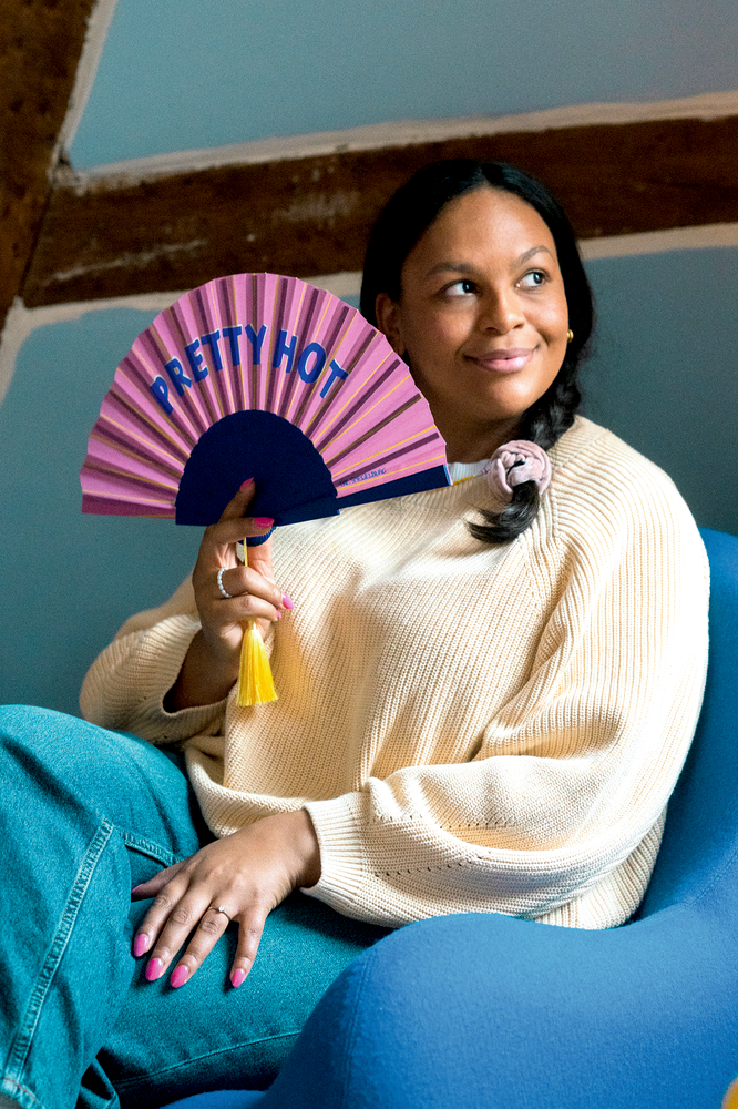Eine Frau mit einem cremefarbenen Pullover hält einen pinken Fächer mit der Aufschrift PRETTY HOT und sitzt auf einem blauen Sessel.