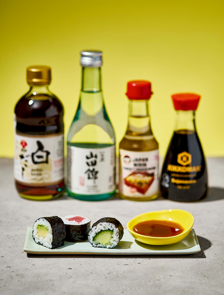 Zwei Sushi-Rollen auf einem Teller mit Sojasauce, davor Flaschen mit Sojasauce, Sake und Gewürzen.