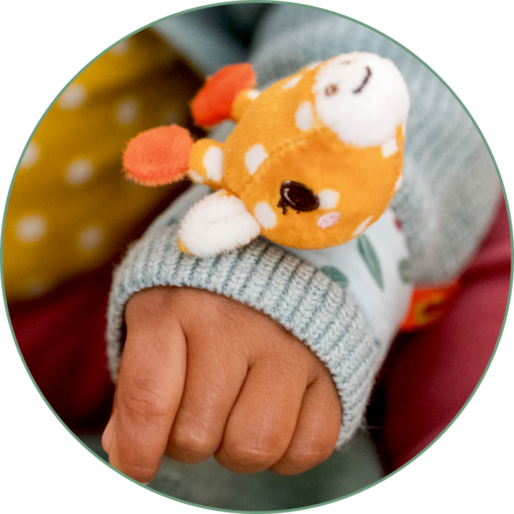 Eine Kinderhand im blauen Ärmel trägt ein plüschiges orangefarbenes Giraffen-Armband.