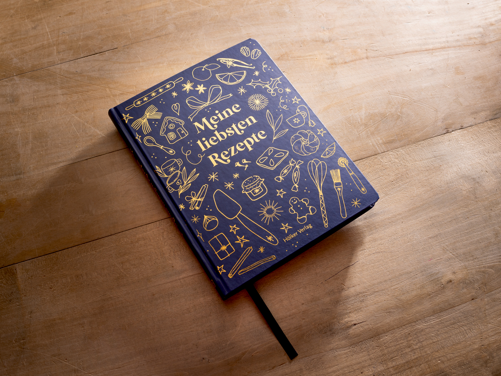 Dunkelblaues Rezeptbuch mit goldenen Küchenutensilien-Motiven und deutschem Text liegt auf einem Holztisch.