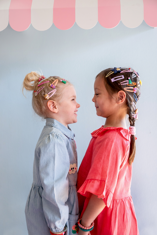 Zwei Kinder stehen sich gegenüber, mit bunten Haarspangen im Haar und farbigen Kleidern in Blau und Rosa.