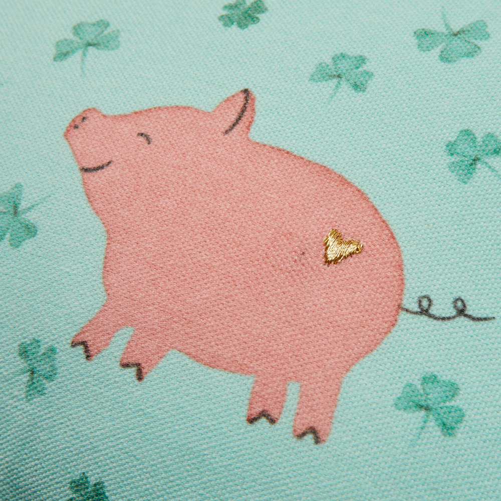 Lächelndes rosa Schwein mit einem goldenen Herz an der Seite, umgeben von grünen Kleeblättern auf blauem Stoff.