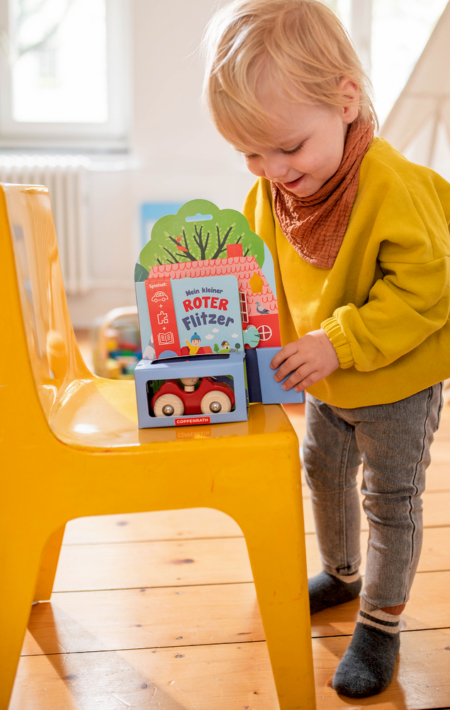 Kleinkind im gelben Pullover spielt drinnen neben einem leuchtend gelben Stuhl mit einem Spielzeugauto.