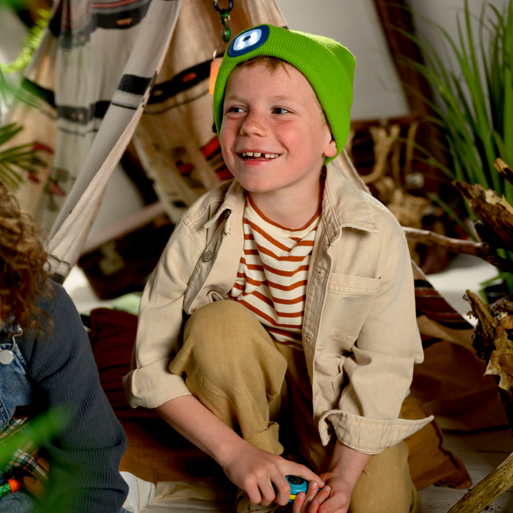 Lächelnder Junge mit grüner Mütze und gestreiftem Hemd sitzt neben einem Zelt, umgeben von Pflanzen.