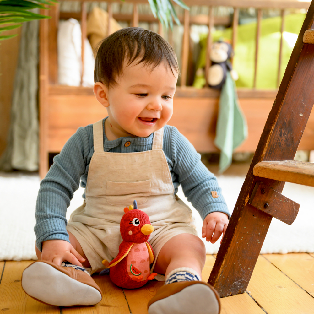 Ein lächelndes Baby in Latzhose sitzt auf dem Boden und hält einen roten Plüschvogel neben einer Holzleiter.