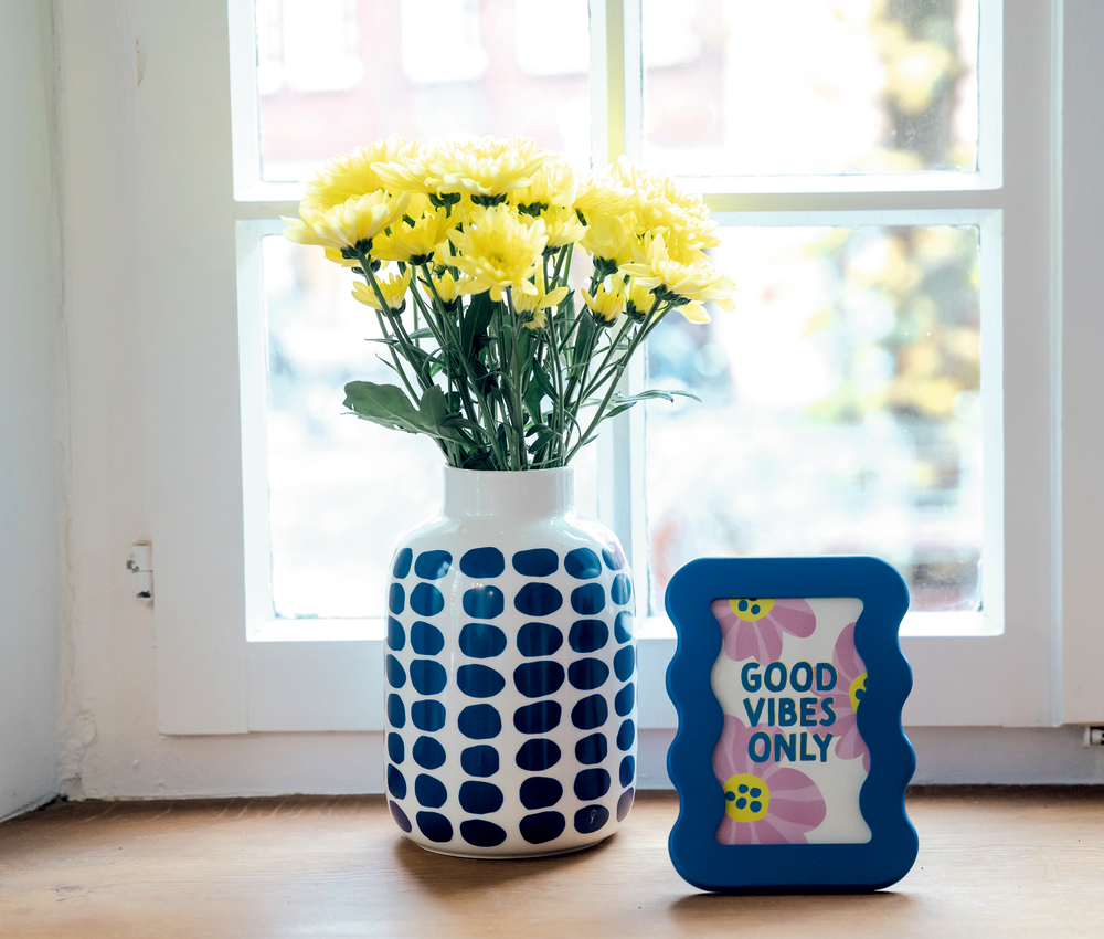 Gelbe Blumen in einer gepunkteten Vase und ein gerahmtes Good Vibes Only Schild stehen auf einem Fensterbrett.