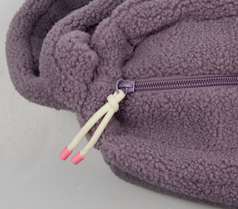 Nahaufnahme einer lila Fleecetasche mit Reißverschluss und zwei cremefarbenen Kordeln mit rosa Spitze.